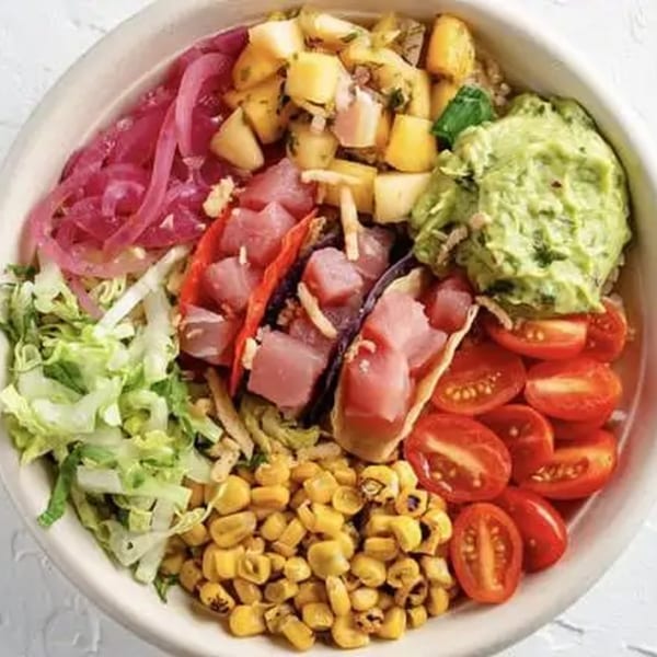 A colorful poke bowl with tuna, pineapple, guacamole, corn, tomatoes, pickled onions, and lettuce.