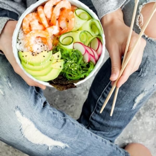 A person in ripped jeans holds a Poke Mahi poke bowl with shrimp, avocado, cucumber, and radish.