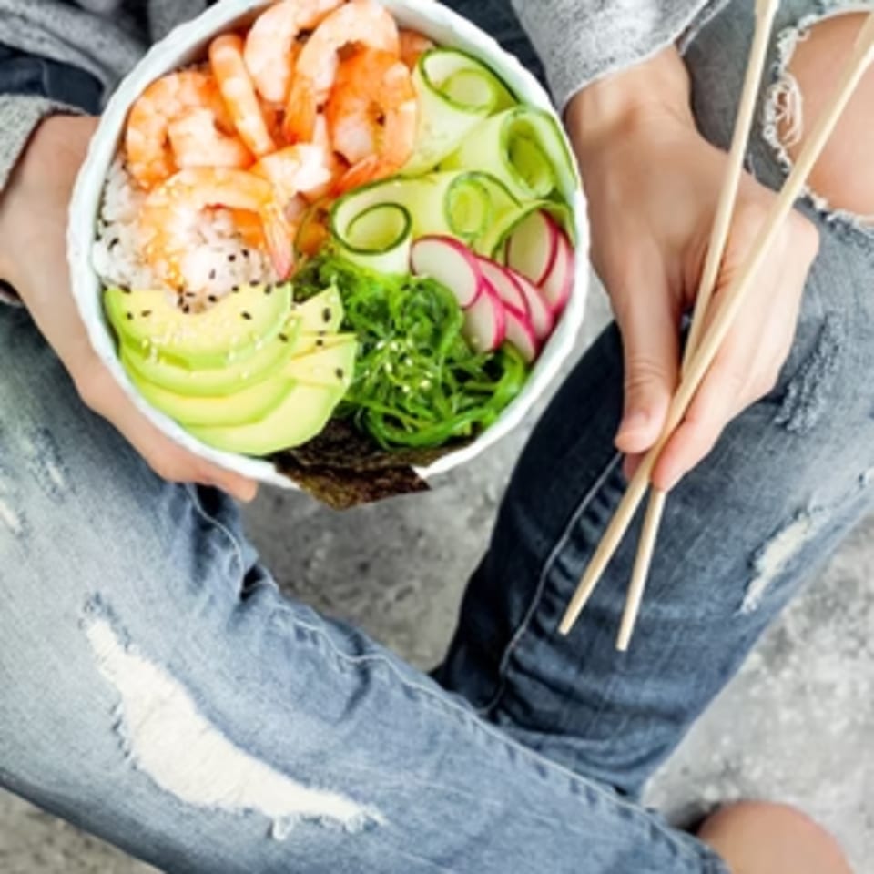 A person in ripped jeans holds a Poke Mahi poke bowl with shrimp, avocado, cucumber, and radish.