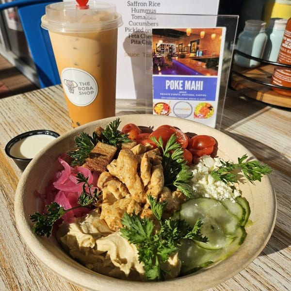 A Poke Mahi bowl with chicken, hummus, vegetables, and a boba tea on a wooden table.